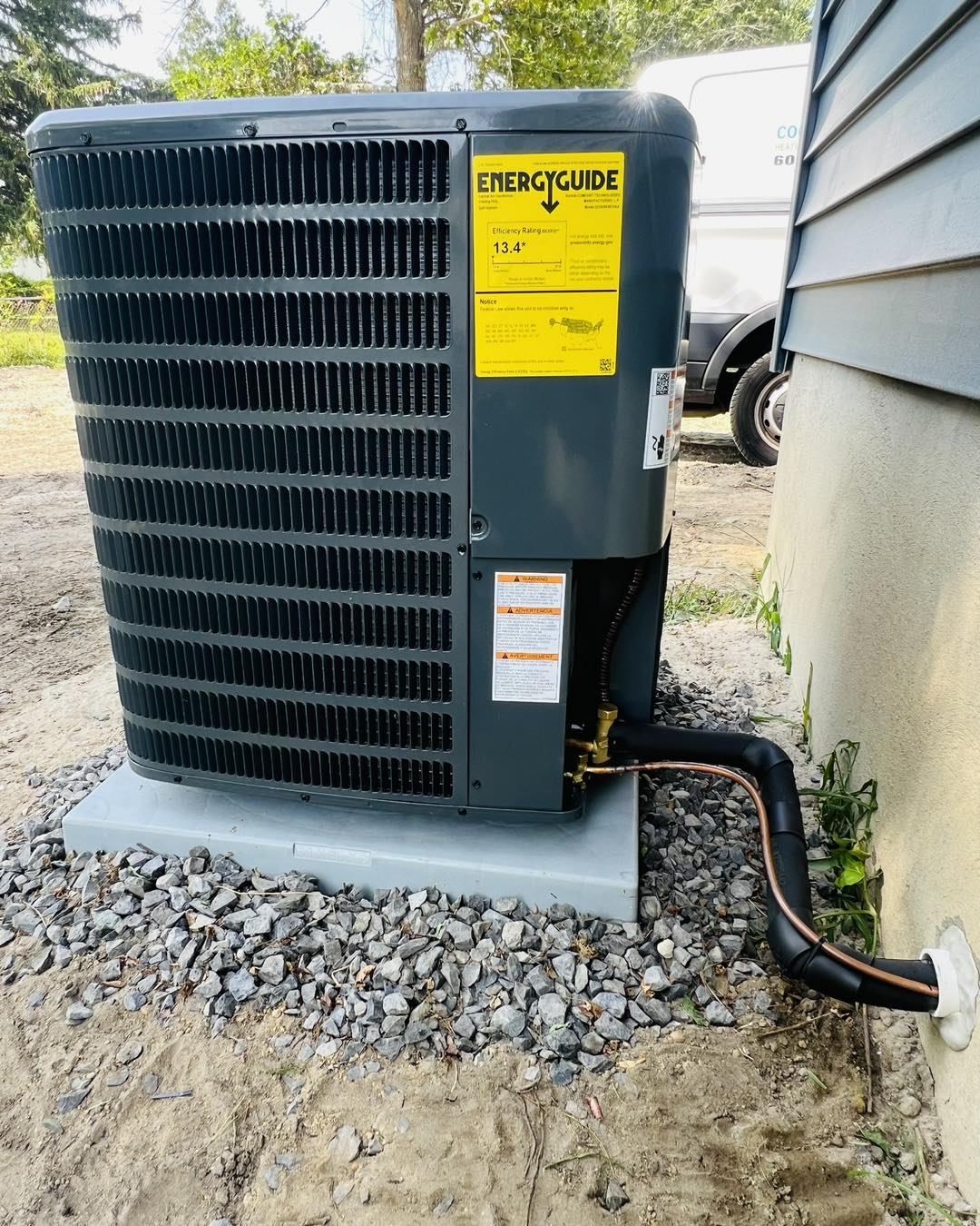 An outdoor air conditioning unit on a concrete pad with an EnergyGuide label, surrounded by gravel and a nearby building wall.