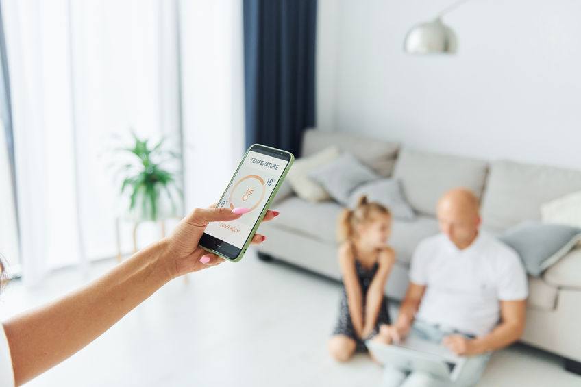 Person holding a smartphone displaying a thermometer app, with a man and child sitting on a couch in the background.