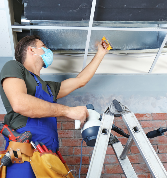 Worker in a blue apron, using a scraper on a ventilation duct while standing on a ladder.