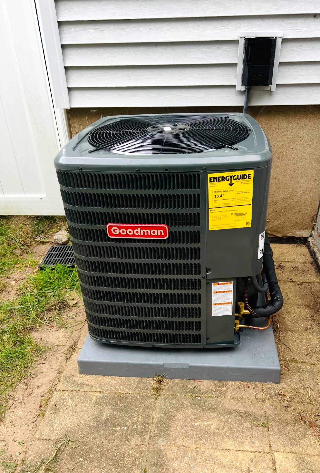 Outdoor Goodman air conditioning unit on a concrete base next to a house exterior wall with a white fence nearby.