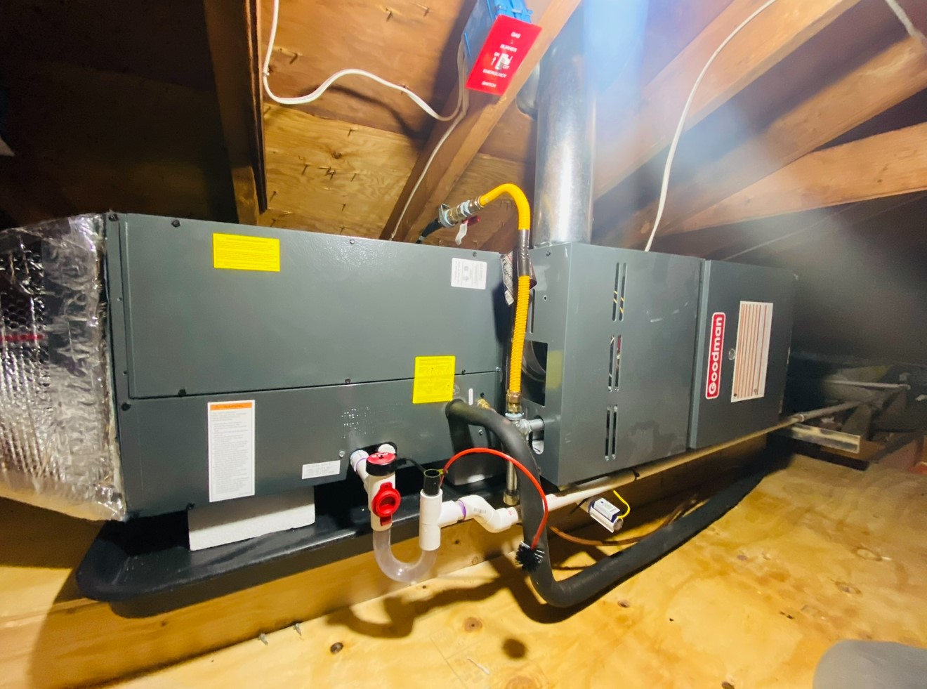 Home attic with an installed Goodman HVAC system, featuring visible ductwork, wiring, and metal vent pipe.